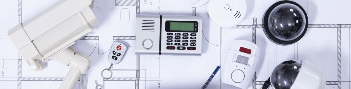 Security devices including CCTV cameras, a keypad, a smoke detector, a remote control, and a motion sensor are arranged on top of architectural blueprints, highlighting the importance of security and safety in building design. by Standex Detect